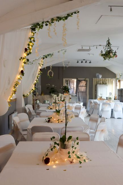 An elegantly decorated event space featuring white round tables draped with crisp tablecloths, each adorned with minimalist floral arrangements in glass vases, predominantly white with hints of green 