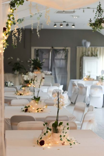 A floral arrangement featuring a tall, slender clear glass vase holding a single white flower, surrounded by delicate white blossoms and dark red roses, with faux fairy lights woven around the base. T