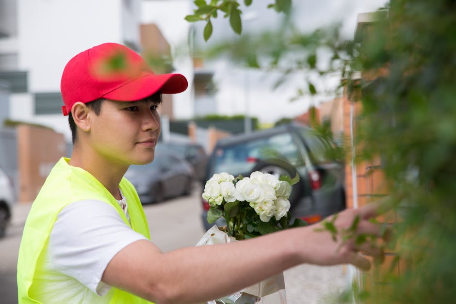 A young male delivery person wearing a bright yellow vest and red cap is outdoors, placing a floral gift against a brick wall surrounded by green foliage. He is holding a bouquet of white hydrangeas w