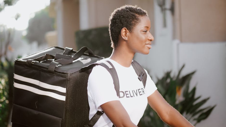 A young woman with short, curly hair and medium-dark skin is outdoors in natural sunlight, carrying a black insulated delivery bag with white stripes on her shoulder. She is dressed in a white T-shirt