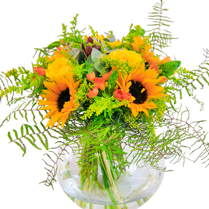 From the first view, this bouquet exudes sunlit vitality-a celebration of the golden afternoons that color Dartford in high summer. Stately sunflowers and soft yellow roses gather in a harmonious embrace, their generous petals catching the light to create a scene of cheerful exuberance. Tucked between are glossy, scarlet hypericum berries, their sheen highlighting the matte textures of rose and sunflower. Cascading ferns and airy sprigs of light green filler spills over the edge, reminiscent of the lush growth you might find winding along the Darent Valley paths. Eucalyptus leaves, with a silvery-blue hue, glimmer at the edge, reinforcing the arrangement's natural, slightly wild charm. The glass vase sits like a transparent pond, cradling the bouquet and revealing each green stem as it dips into fresh water. Up close, the arrangement offers a gentle sensory mix: the sunflowers' suede softness, the dense spirals of rose petals, the fragrant freshness of newly cut stems. This design is perfect for life's happiest milestones or as a daily reminder of nature's abundance-ideal for homes and celebrations around Dartford, where flowers speak the language of joy.