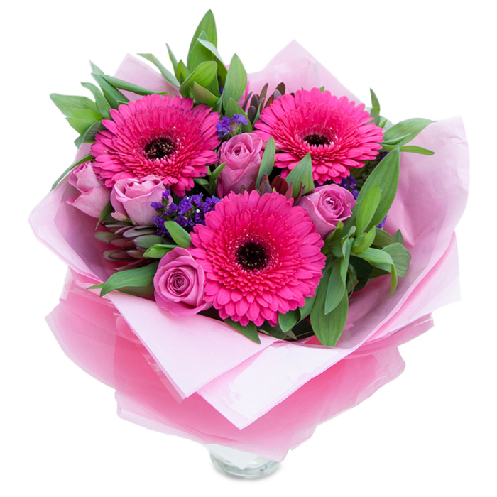 I always think that the charm of fresh pink flowers lies in their ability to balance playfulness with quiet grace, and this bouquet captures that feeling for anyone receiving blooms in and around Dartford. Three wide-eyed gerbera daisies unfurl in a vivid fuchsia, their radiant petals arranged like small, joyful suns with dark centres that anchor the colour. Nestled among them are rosebuds in a muted lavender-pink or dusty rose - petals still tightly curled, creating soft, sculptural forms that invite touch. Tiny clusters of purple statice are tucked throughout, their papery texture adding a delicate blue-violet punctuation that lifts the composition and recalls wildflowers drifting through a garden patch. Ample greenery - broad, glossy leaves suggesting salal and the silvery hint of eucalyptus - frames the flowers and adds a cool, dewy counterpoint to the warm pinks. The bouquet is finished with an inner sleeve of pale pink tissue and an outer wrap of translucent fuchsia-pink cellophane, slightly crinkled so it catches the light in a subtle shimmer; the careful folds and the visible rim of a clear glass vase beneath show it has been prepared for immediate display. There's a gentle scent of rose softened by the green sweetness of gerbera stems, and the tactile mix of velvety petals, crisp foliage, and crinkled wrap creates an intimate gift experience. Thoughtful for Mother's Day, birthdays, or simply to brighten someone's day, this arrangement travels well across Dartford streets from Joyden's Wood to the area near Bluewater, offering a refined, cheerful presence wherever it arrives.